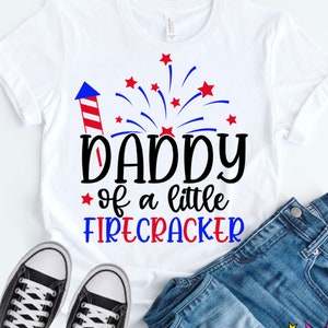 May include: A white t-shirt with a red, white, and blue design that says "Daddy of a little Firecracker". The design includes a firework and a rocket. The shirt is on a blue denim background with a pair of white sneakers and the text "SVG + DXF + EPS + PNG + JPG" at the bottom.
