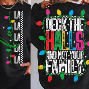 Puede incluir: Sudadera negra con diseño de luces navideñas. La parte delantera presenta la palabra "LA" repetida con una cadena de luces. La parte trasera dice "Deck the Halls and Not Your Family" con luces de colores.
