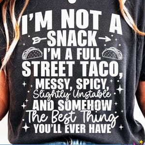 Op de afbeelding: Donkergrijs t-shirt met witte tekst: "I'M NOT A SNACK - I'M A FULL STREET TACO, MESSY, SPICY, Slightly Unstable AND SOMEHOW The Best Thing YOU'LL EVER HAVE." Met taco- en pijlgraphics. SVG, PNG, EPS, DXF, JPG formaten.