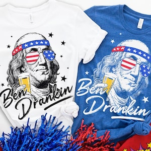 May include: White and blue t-shirts with a graphic of Benjamin Franklin wearing sunglasses with an American flag design and a headband. The text "Ben Drankin" is below the image. Black stars are scattered around the image.