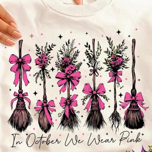 Op de afbeelding: Wit sweatshirt met een ontwerp van vijf decoratieve heksenbezems, elk versierd met roze strikken en bloemstukken. De tekst "In October We Wear Pink" staat eronder. Het ontwerp bevat zwarte sterren en roze accenten.