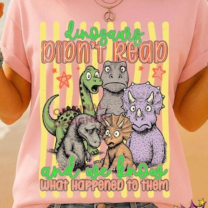 May include: Pink t-shirt featuring a colorful graphic of five cartoon dinosaurs with the text "Dinosaurs Didn't Read and We Know What Happened to Them." The design includes stars and vertical stripes.