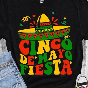 Peut inclure: Un t-shirt noir avec un motif graphique coloré représentant un sombrero, le texte "CINCO DE MAYO FIESTA" dans une police rétro et des étoiles. Le motif est dans les tons de rouge, jaune, vert et noir.