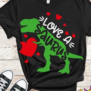 Może przedstawiać: Czarny T-shirt z zielonym dinozaurem trzymającym czerwone serce i tekstem "=LOVE A= Saurus".