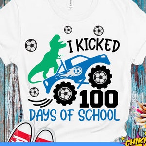 Op de afbeelding: Een wit T-shirt met een blauwe monstertruck, een groene dinosaurus en voetballen. De tekst op het shirt luidt "Ik heb 100 dagen school getrapt".