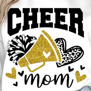 Puede incluir: Sudadera blanca con un diseño en negro y dorado. El diseño incluye la palabra "CHEER" en letras negras en negrita, un megáfono dorado con purpurina, un pompón en blanco y negro, un corazón con estampado de leopardo y la palabra "mamá".