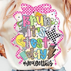 May include: Cream-colored t-shirt with a colorful graphic that reads "Bruh It's Test Day" with various design elements. The design includes a pink bow, stars, lightning bolts, and the hashtag #yougotthis.