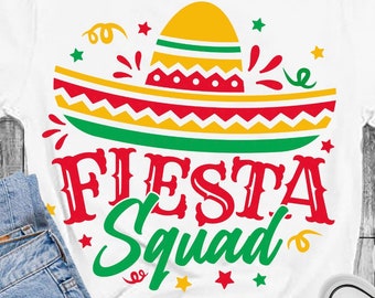 May include: A white t-shirt with a green, yellow, and red sombrero design and the text "Fiesta Squad" in green and red. The shirt is on a wooden surface with a pair of black and white sneakers and a pair of blue jeans.