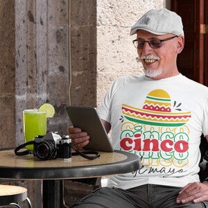 Peut inclure: T-shirt blanc avec un motif de sombrero color&eacute; et l'inscription "Cinco de Mayo". L'image montre &eacute;galement un appareil photo vintage, une boisson verte avec une tranche de citron vert et une personne portant un chapeau et des lunettes.