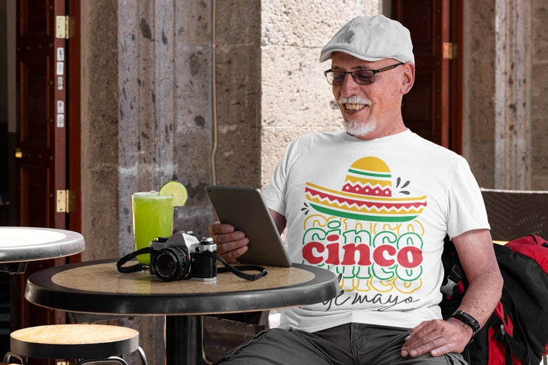 Peut inclure: T-shirt blanc avec un motif de sombrero color&eacute; et l'inscription "Cinco de Mayo". L'image montre &eacute;galement un appareil photo vintage, une boisson verte avec une tranche de citron vert et une personne portant un chapeau et des lunettes.