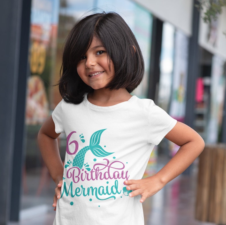 May include: White t-shirt with a colorful mermaid-themed design. The graphic includes the number "6", a mermaid tail, and the words "Birthday Mermaid" in a script font. The shirt is worn by a young girl.