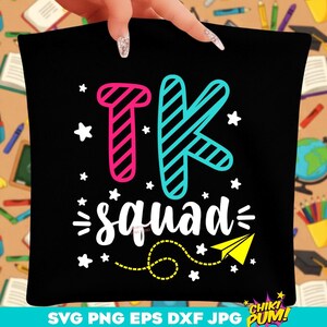 May include: Black pillow with the text "TK squad" in white, with pink and blue striped letters. A yellow paper airplane and white stars decorate the design. The pillow is held by a hand.