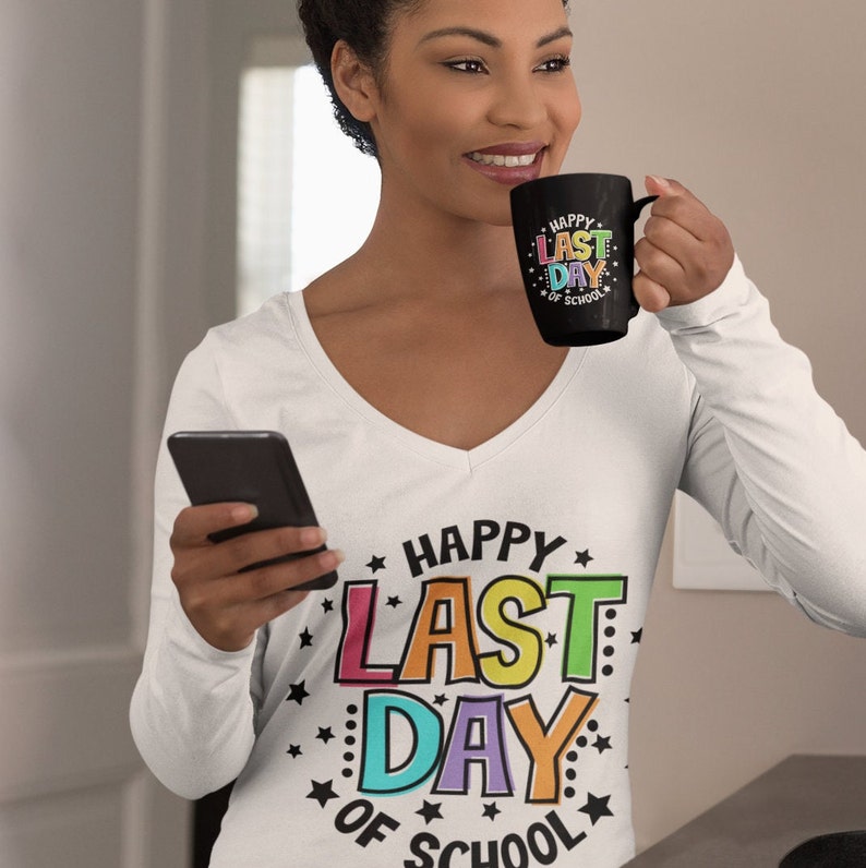Puede incluir: Una camiseta blanca de manga larga con un gr&aacute;fico colorido que dice "Happy Last Day of School" rodeado de estrellas. La persona de la imagen sostiene una taza negra con el mismo gr&aacute;fico.