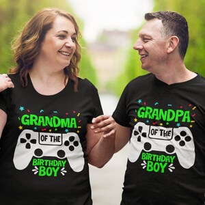 May include: Two people wearing black t-shirts with a white video game controller graphic. The text on the shirts reads "Grandma of the Birthday Boy" and "Grandpa of the Birthday Boy".