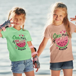 May include: Two girls wearing matching t-shirts with a watermelon graphic and the text "One in a Melon Sister". The girl on the left is wearing a green shirt and the girl on the right is wearing a pink shirt. They are both smiling and holding hands.