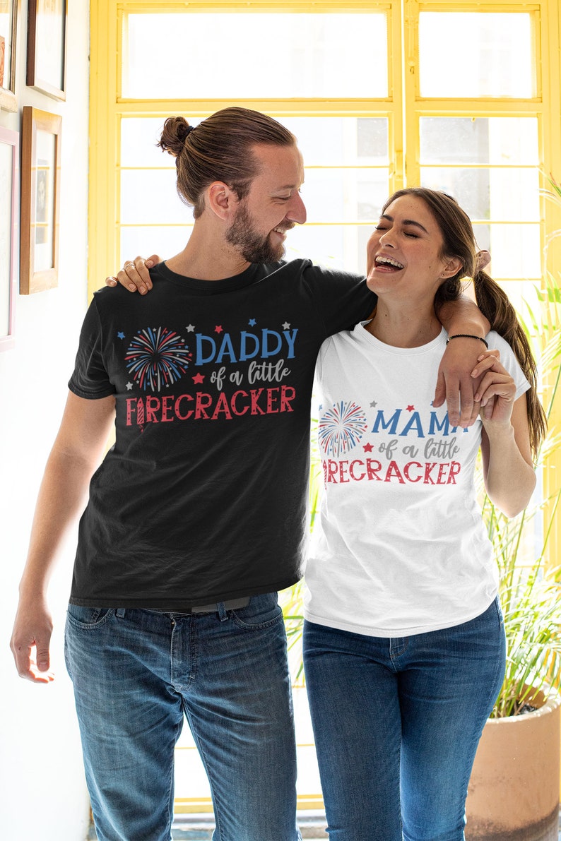 Op de afbeelding: Een stel draagt ​​bijpassende zwarte en witte T-shirts met rood-wit-blauwe vuurwerkgrafieken. De tekst op de shirts luidt "Daddy of a little Firecracker" en "Mama of a little Firecracker".