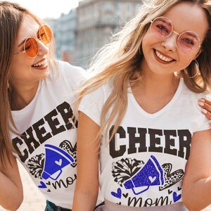 May include: Two people wearing white t-shirts with the text "Cheer Mom" in black and blue glitter. The shirts feature a blue megaphone with a white pom-pom and hearts.