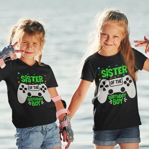 May include: Two children wearing black t-shirts with a white video game controller graphic and the text "Sister of the Birthday Boy". The shirts have colorful confetti designs. The children are standing outdoors in front of a body of water.