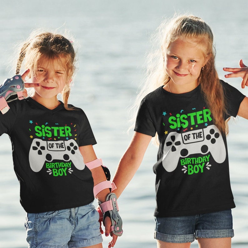 May include: Two children wearing black t-shirts with a white video game controller graphic and the text "Sister of the Birthday Boy". The shirts have colorful confetti designs. The children are standing outdoors in front of a body of water.