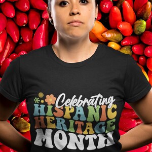 Pode incluir: Uma pessoa vestindo uma camiseta preta com o texto "Celebrating Hispanic Heritage Month" em letras retr&ocirc; coloridas. A pessoa est&aacute; em p&eacute; em frente a um fundo de pimentas vermelhas.