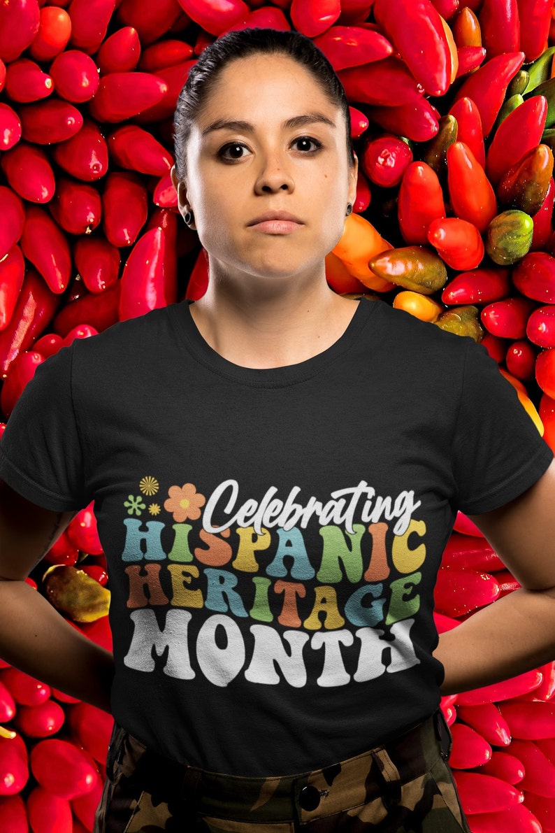 Pode incluir: Uma pessoa vestindo uma camiseta preta com o texto "Celebrating Hispanic Heritage Month" em letras retr&ocirc; coloridas. A pessoa est&aacute; em p&eacute; em frente a um fundo de pimentas vermelhas.