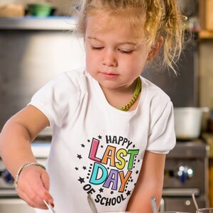 Puede incluir: Una ni&ntilde;a peque&ntilde;a con una camiseta blanca con un gr&aacute;fico colorido que dice "Happy Last Day of School" est&aacute; mirando algo en sus manos.