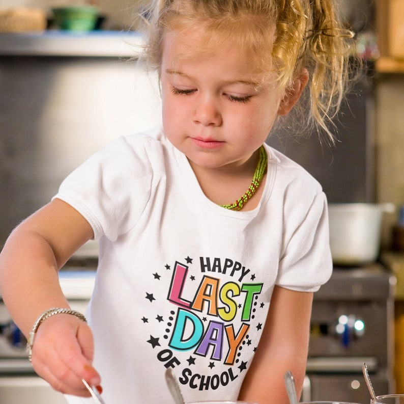Puede incluir: Una ni&ntilde;a peque&ntilde;a con una camiseta blanca con un gr&aacute;fico colorido que dice "Happy Last Day of School" est&aacute; mirando algo en sus manos.