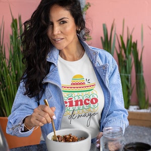 Peut inclure: Une femme porte un t-shirt blanc avec un motif Cinco de Mayo, une veste en jean et tient un bol de nourriture. Le t-shirt pr&eacute;sente un sombrero et les mots "Cinco de Mayo" en rouge, jaune et vert. Un bocal en verre et une tasse noire sont sur la table.