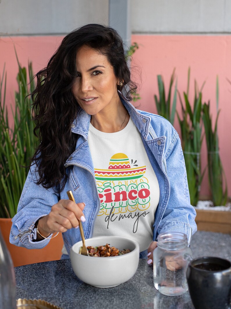Peut inclure: Une femme porte un t-shirt blanc avec un motif Cinco de Mayo, une veste en jean et tient un bol de nourriture. Le t-shirt pr&eacute;sente un sombrero et les mots "Cinco de Mayo" en rouge, jaune et vert. Un bocal en verre et une tasse noire sont sur la table.