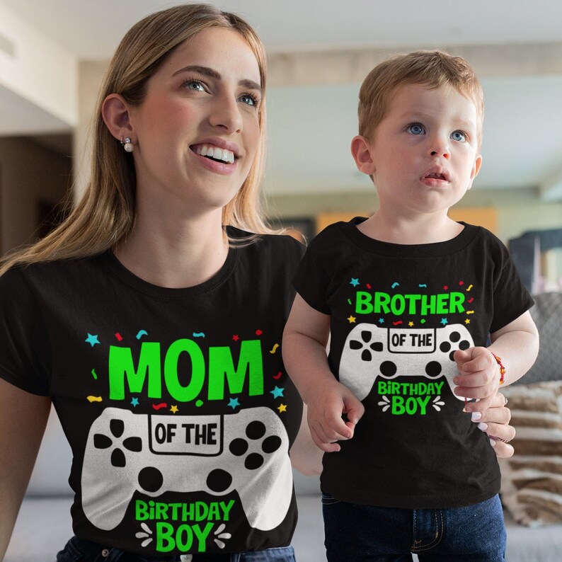 May include: A black t-shirt with a green and white video game controller graphic. The text "MOM OF THE BIRTHDAY BOY" is printed on the shirt. A smaller black t-shirt with a green and white video game controller graphic and the text "BROTHER OF THE BIRTHDAY BOY" is printed on the shirt.