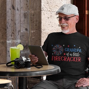 Op de afbeelding: Een man draagt een zwart T-shirt met een rood, wit en blauw vuurwerkmotief en de tekst "Grandpa of a little firecracker". Hij zit aan een tafel buiten, met een grijze pet en een bril op.