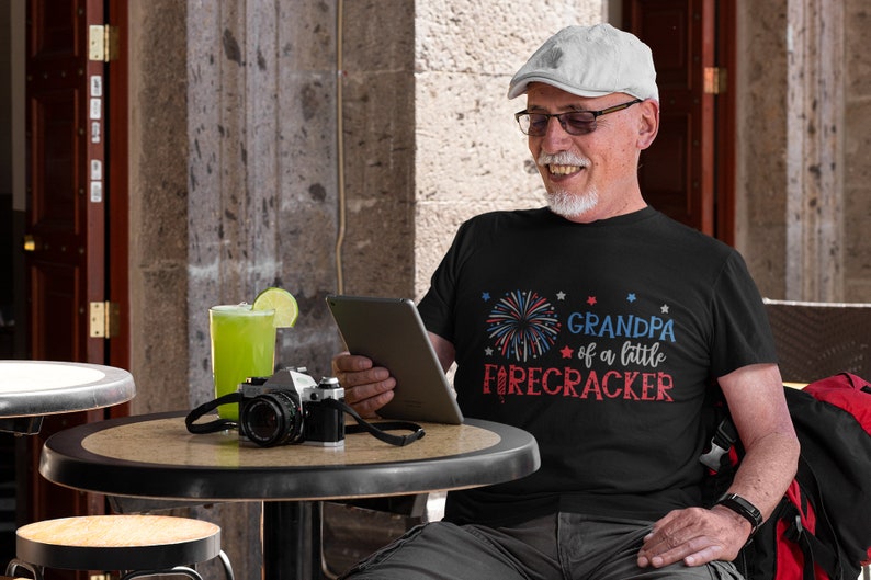 Op de afbeelding: Een man draagt een zwart T-shirt met een rood, wit en blauw vuurwerkmotief en de tekst "Grandpa of a little firecracker". Hij zit aan een tafel buiten, met een grijze pet en een bril op.