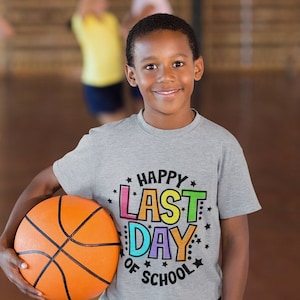 Puede incluir: Una persona joven con una camiseta gris con el texto "Happy Last Day of School" en letras de colores sostiene un bal&oacute;n de baloncesto naranja.