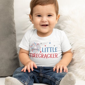 Op de afbeelding: Een witte baby romper met een rood, wit en blauwe vuurwerk afbeelding en de tekst "LITTLE FIRECRACKER". Een baby draagt de romper en zit op een witte bank.