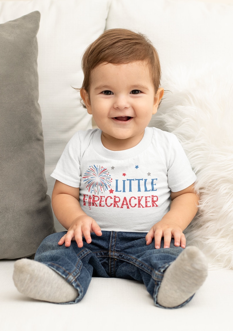 Op de afbeelding: Een witte baby romper met een rood, wit en blauwe vuurwerk afbeelding en de tekst "LITTLE FIRECRACKER". Een baby draagt de romper en zit op een witte bank.