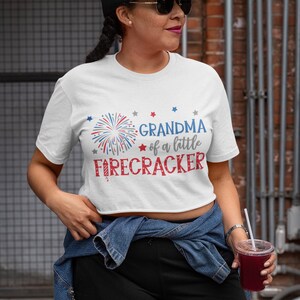 Op de afbeelding: Wit T-shirt met een rood-wit-blauwe afbeelding van vuurwerk en de tekst "Grandma of a little firecracker".