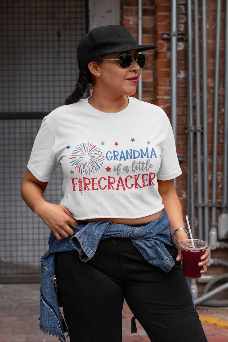 Op de afbeelding: Wit T-shirt met een rood-wit-blauwe afbeelding van vuurwerk en de tekst "Grandma of a little firecracker".