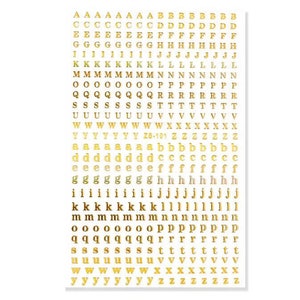 May include: Gold metallic alphabet letter stickers on a white background. The stickers include uppercase and lowercase letters, numbers, and symbols. The letters are in a sans-serif font and are arranged in rows and columns.
