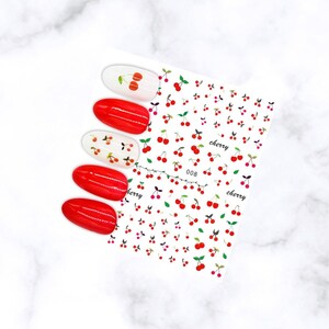 May include: Nail art stickers featuring a cherry design. The sheet has a repeating pattern of red cherries with green leaves and the word "cherry". Four red painted nails and two white nails with cherry designs are also shown.