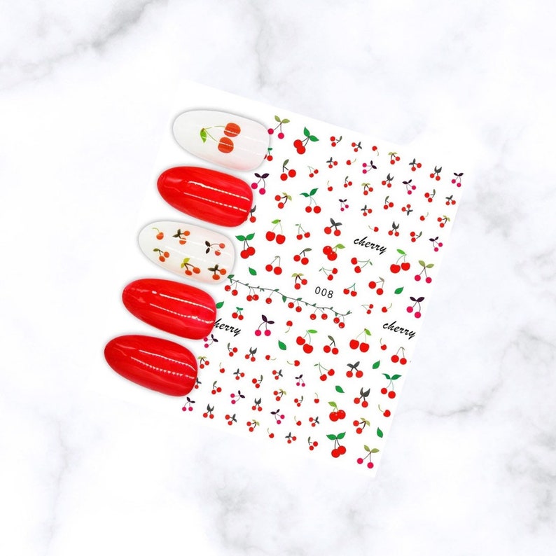 May include: Nail art stickers featuring a cherry design. The sheet has a repeating pattern of red cherries with green leaves and the word "cherry". Four red painted nails and two white nails with cherry designs are also shown.