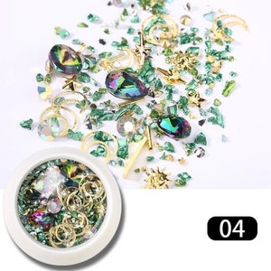 May include: A collection of nail art supplies featuring iridescent teardrop-shaped gems, gold star and moon charms, and green crushed glass. A white container holds similar items. The number "04" is in a black rectangle.
