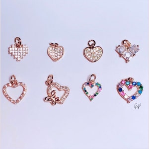 May include: Collection of heart-shaped charms in various designs. Some are encrusted with clear crystals, others feature colorful gemstones, and one has a butterfly accent. All are gold-toned with a loop for attaching to a necklace or bracelet.