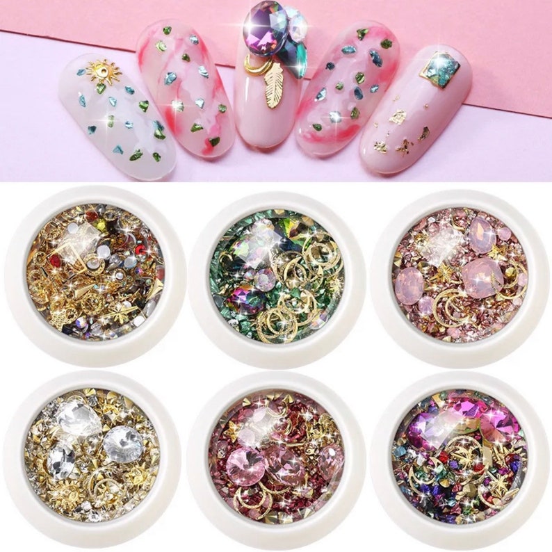 May include: A collection of nail art supplies, including various rhinestones, gems, and gold-colored metal charms. The image also shows several manicured nails with decorative elements, including glitter, gems, and gold leaf.