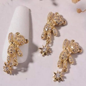 K&ouml;nnte beinhalten: Goldfarbene Nagelkunst-Dekorationen. Drei Teddyb&auml;r-Anh&auml;nger, jeder mit winzigen, klaren Kristallen besetzt, h&auml;ngen an sternf&ouml;rmigen Anh&auml;ngern. Ein Anh&auml;nger befindet sich auf einem wei&szlig;en Nagel. Der Hintergrund ist eine weiche, neutrale Farbe.