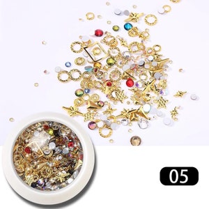 May include: A collection of nail art supplies, including gold-colored metal charms in various shapes like stars, rings, and squares, along with colorful rhinestones and gems. The items are scattered on a white surface, with a small container filled with similar items.