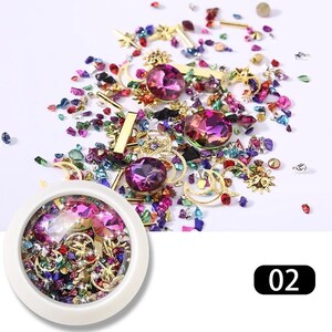 May include: A collection of colorful nail art supplies, including iridescent gems, gold-colored charms, and crushed glass. The assortment features large, faceted purple gems, star and moon charms, and a container filled with similar materials. The number "02" is in the bottom right corner.