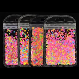 May include: Four clear plastic bags filled with colorful, neon-colored shapes. The shapes include stars, hearts, and small round beads. The bags are sealed and have a small hole at the top for hanging. The background is black.