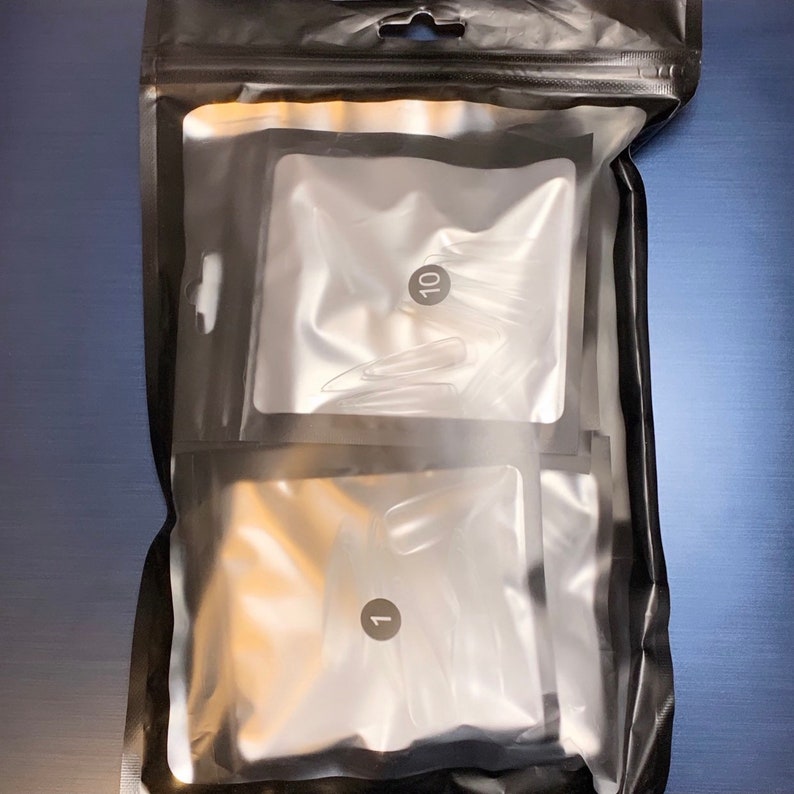 May include: A black plastic bag containing two clear plastic pouches. Each pouch contains clear, pointed nail tips. One pouch is labeled with the number "1" and the other with the number "10". The bag is sealed at the top.