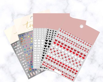 May include: A collection of nail art stickers featuring heart designs in various colors, including red, black, white, and iridescent. Each sheet has a pink header with a hole for hanging. The stickers are arranged on a light gray background.