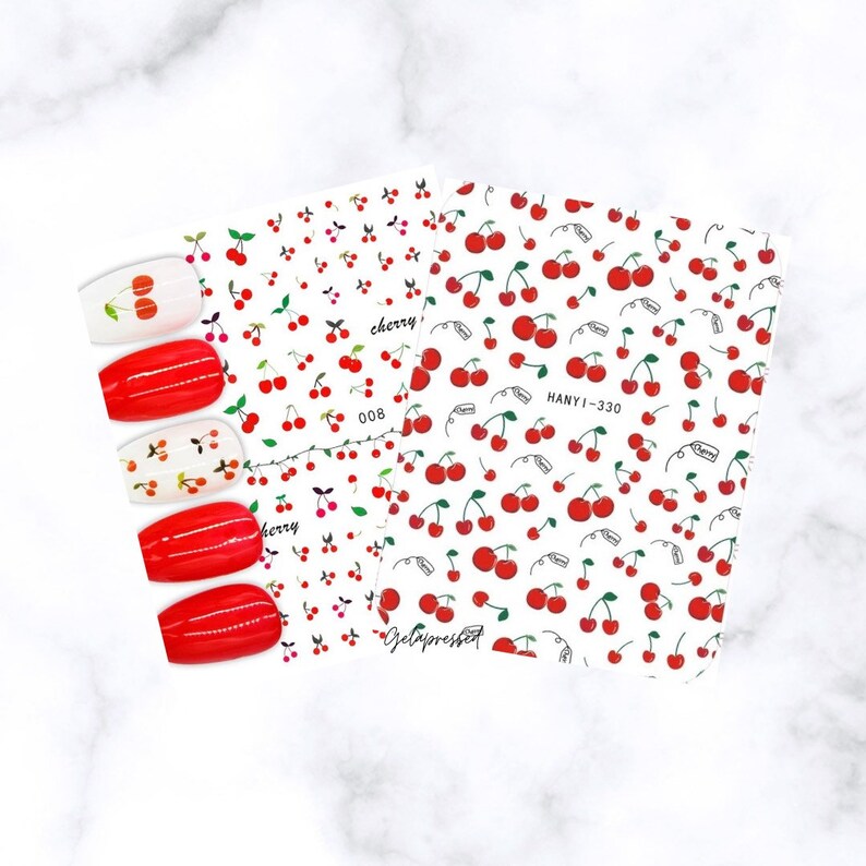 May include: Two sheets of nail art stickers with a cherry design. The stickers feature red cherries with green stems scattered across a white background. Several painted nails are displayed, showcasing the cherry design.